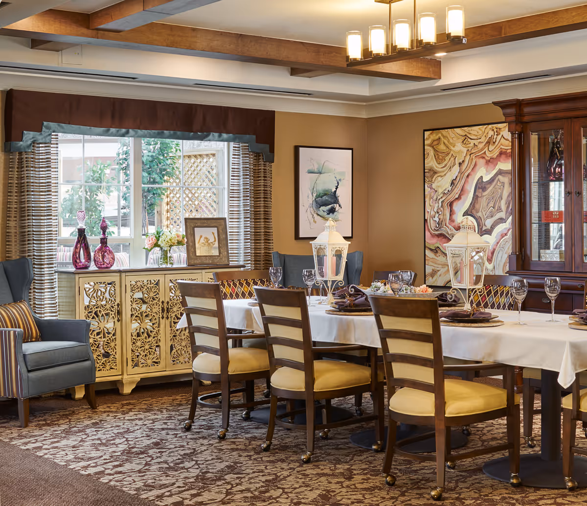 Warmly decorated dining room with a long set table, wooden chairs, decorative cabinets, artwork and a window with valance.