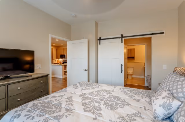 A bedroom with a patterned bedspread on the bed, a dresser with a flat-screen TV on top, and two doors leading to a kitchen area and a bathroom. The bathroom door is a sliding barn-style door, partially open showing a toilet inside.