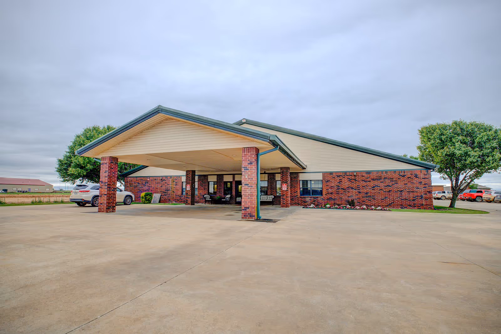 Exterior view of a single-story brick building with a large covered entrance supported by brick columns. There are a few cars parked near the building and some trees around the parking area under a cloudy sky.