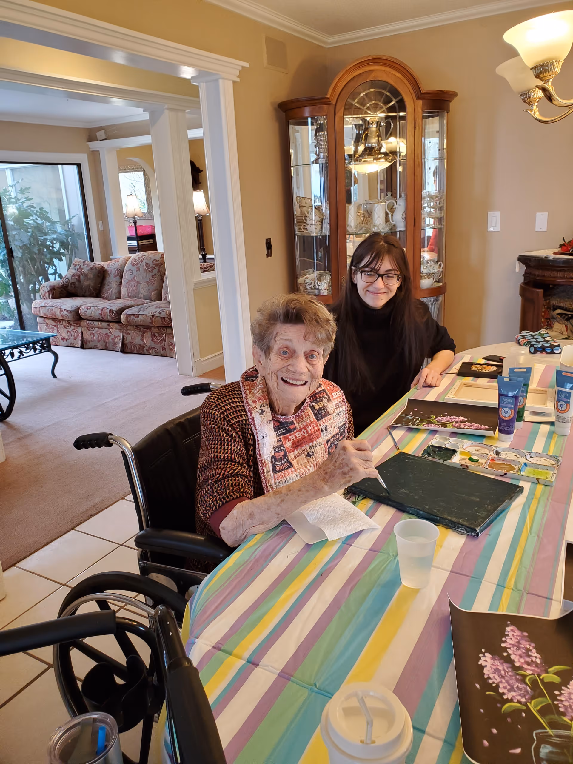 An elderly woman in a wheelchair and a younger woman smile while painting at a table covered with a colorful striped tablecloth in a home-like living room.