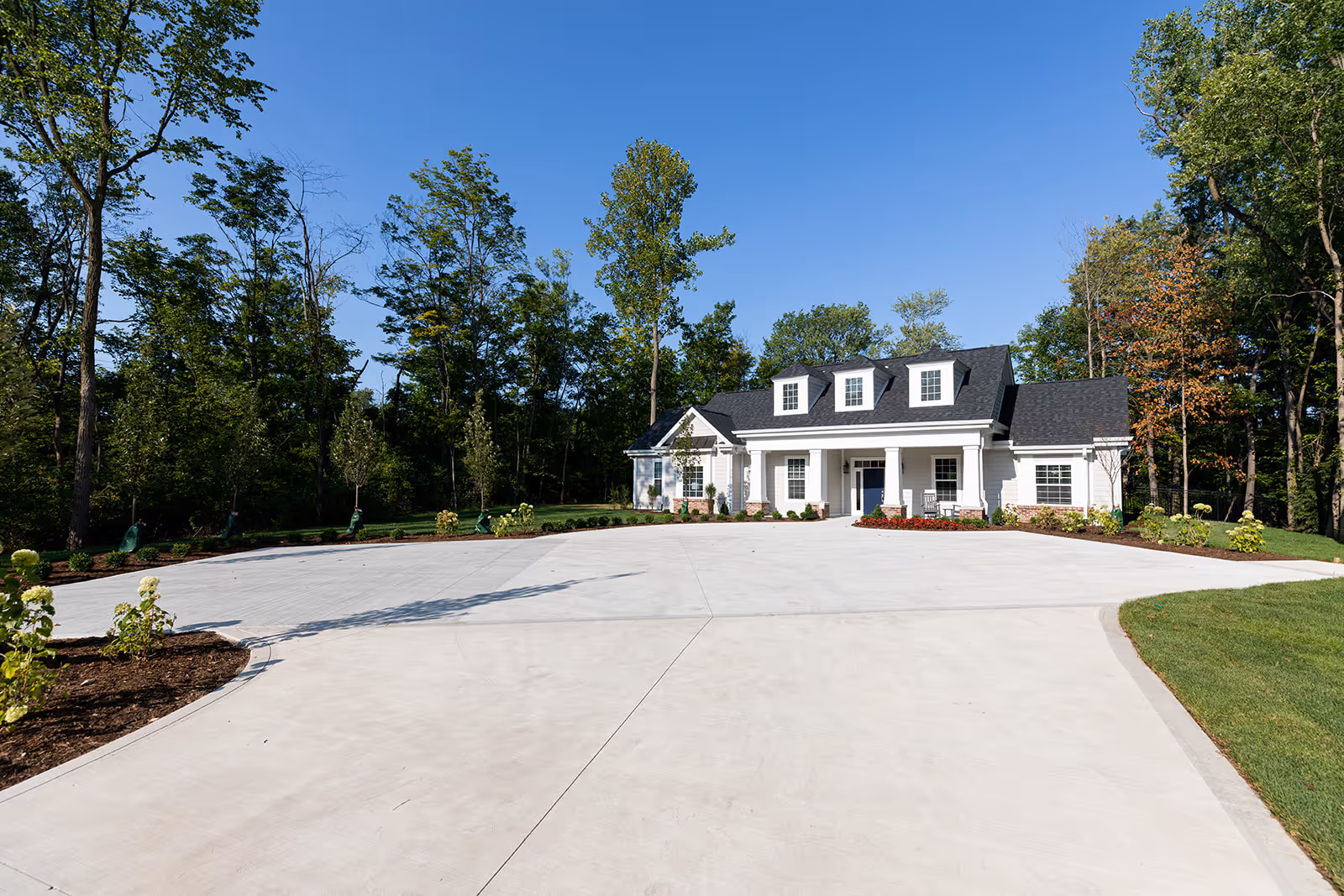A large white single-story building with a dark roof and three dormer windows, surrounded by trees and greenery under a clear blue sky. The building has a wide concrete driveway and landscaped garden beds with small plants and flowers.