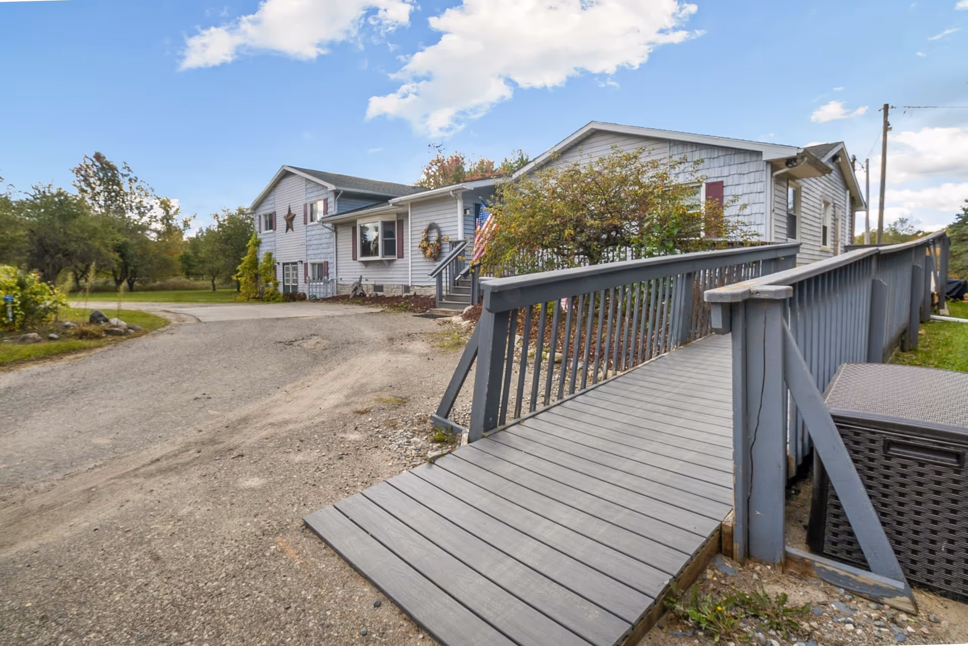 Exterior view of a single-story residential building with gray siding and maroon shutters. There is a wooden ramp with railings leading up to the entrance, which is decorated with a wreath and an American flag. The surrounding area includes a gravel driveway, some bushes, and trees under a partly cloudy sky.