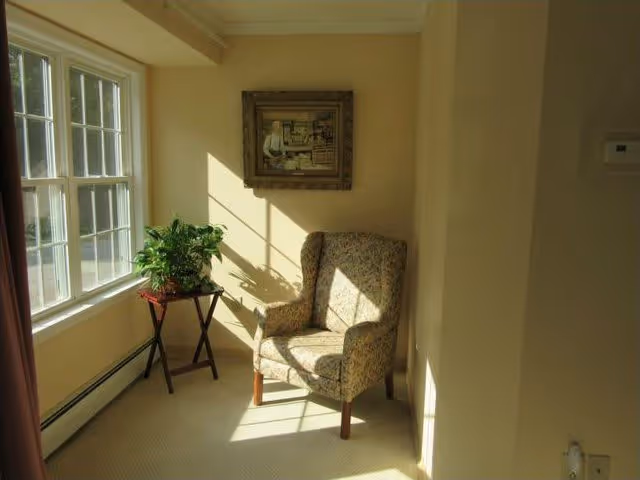 A cozy corner with a floral upholstered armchair positioned near a large window letting in sunlight. Next to the chair is a small wooden table holding a green potted plant. A framed painting hangs on the beige wall above the chair.