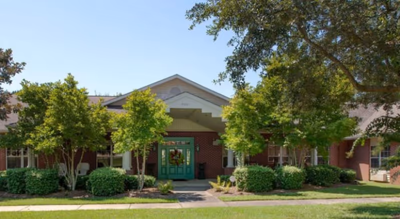 Front entrance of a single-story brick assisted living building with a covered portico, green double doors, and landscaped trees and shrubs.