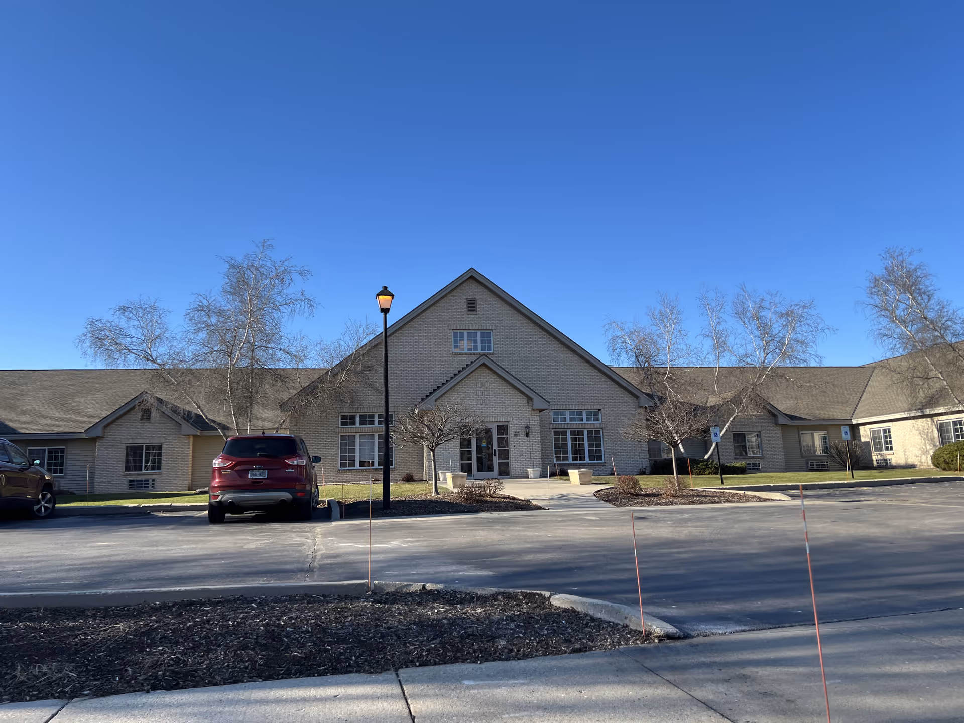 Front exterior view of a single-story senior living facility building with a peaked roof, beige brick facade, several windows, a central entrance with glass doors, a parking lot with a few cars, leafless trees, and a clear blue sky.