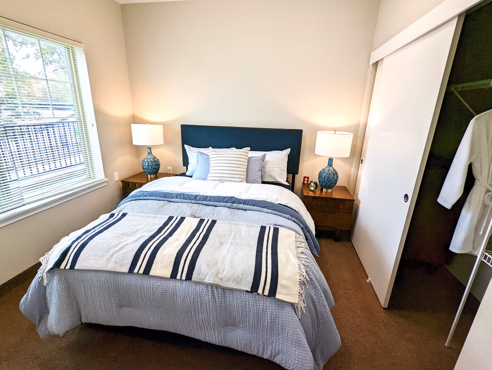 A neatly made bed with a blue headboard, blue and white bedding, and a striped throw blanket in a bedroom. There are two wooden nightstands on either side of the bed, each with a blue lamp. A window with blinds is on the left wall, and a closet with sliding doors and a white robe hanging inside is on the right.
