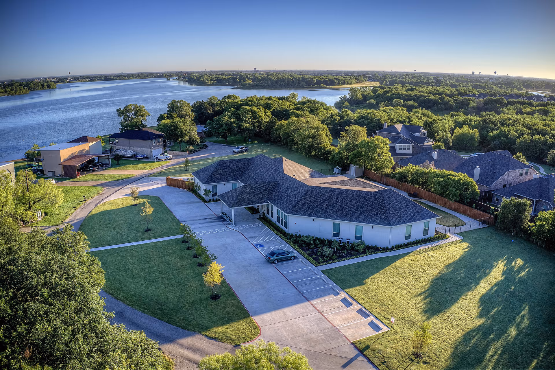 Aerial view of a single-story building with a dark roof surrounded by green lawns and trees, located near a large body of water. The building has a parking area with a few cars and is situated in a peaceful, suburban setting with other houses and dense greenery nearby under a clear blue sky.