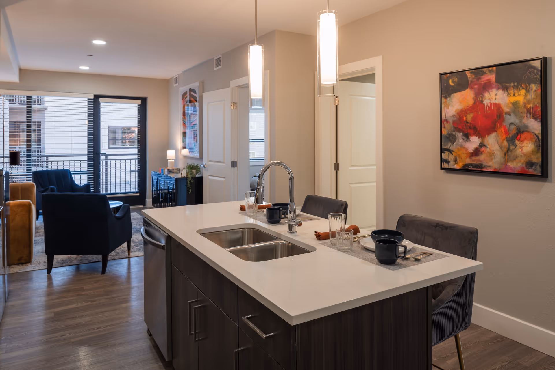 Modern interior of a senior living facility showing a kitchen island with a sink, two place settings with dishes and glasses, and two pendant lights hanging above. In the background, there is a living area with armchairs, a side table with a lamp, and large windows with blinds. The walls are decorated with colorful abstract paintings.