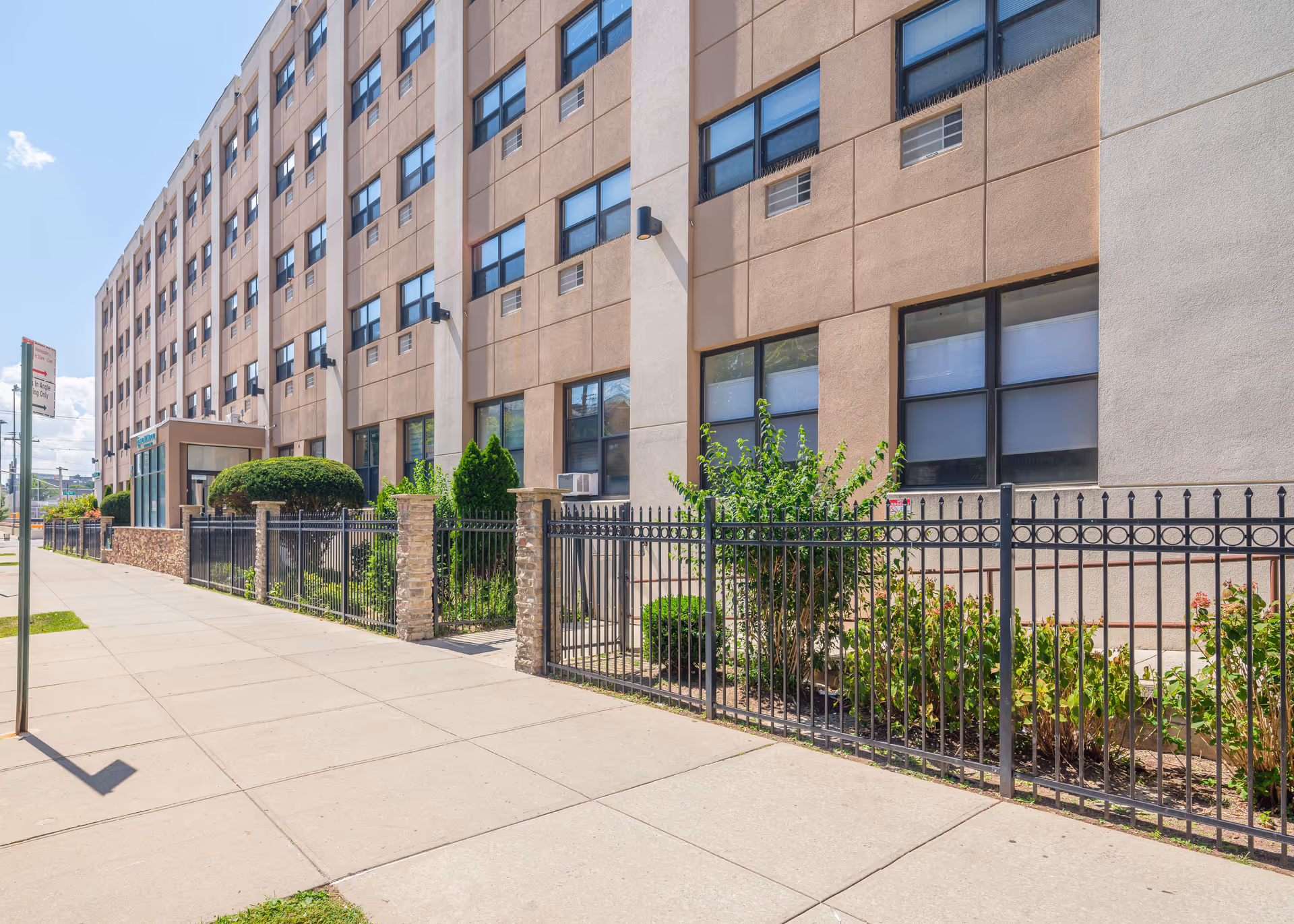 Exterior view of a multi-story senior living facility named Bronxwood with a sidewalk, black metal fence, and landscaped bushes and plants along the building.
