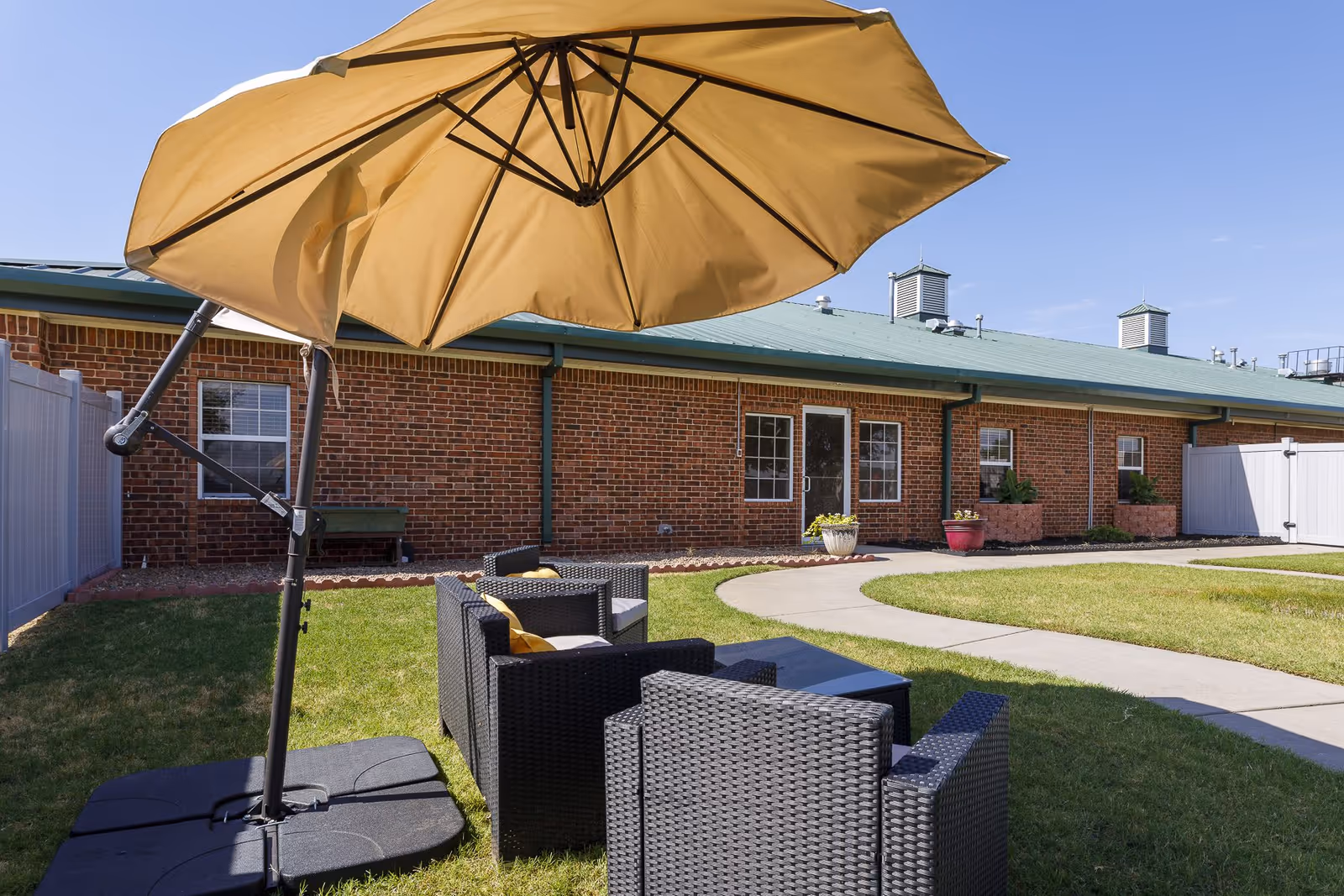 Outdoor seating area with black wicker chairs and a large beige umbrella on a grassy lawn in front of a brick building with green roof and windows.