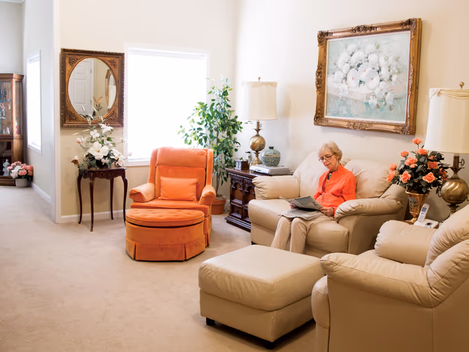 A cozy living room with beige leather seating, an orange armchair, floral decor, and a person reading on the sofa.