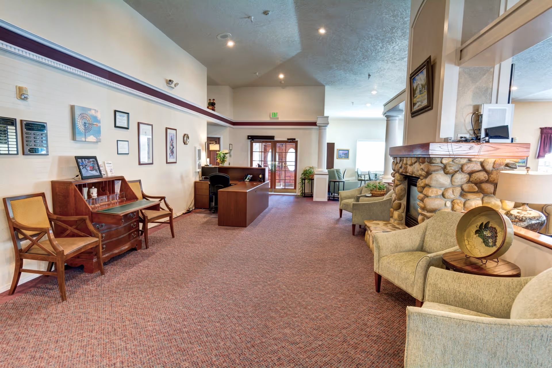 A spacious senior living facility lobby with a reception desk, wooden chairs, framed pictures on the wall, and a stone fireplace surrounded by comfortable armchairs. The area is well-lit with natural light coming through glass doors and windows.