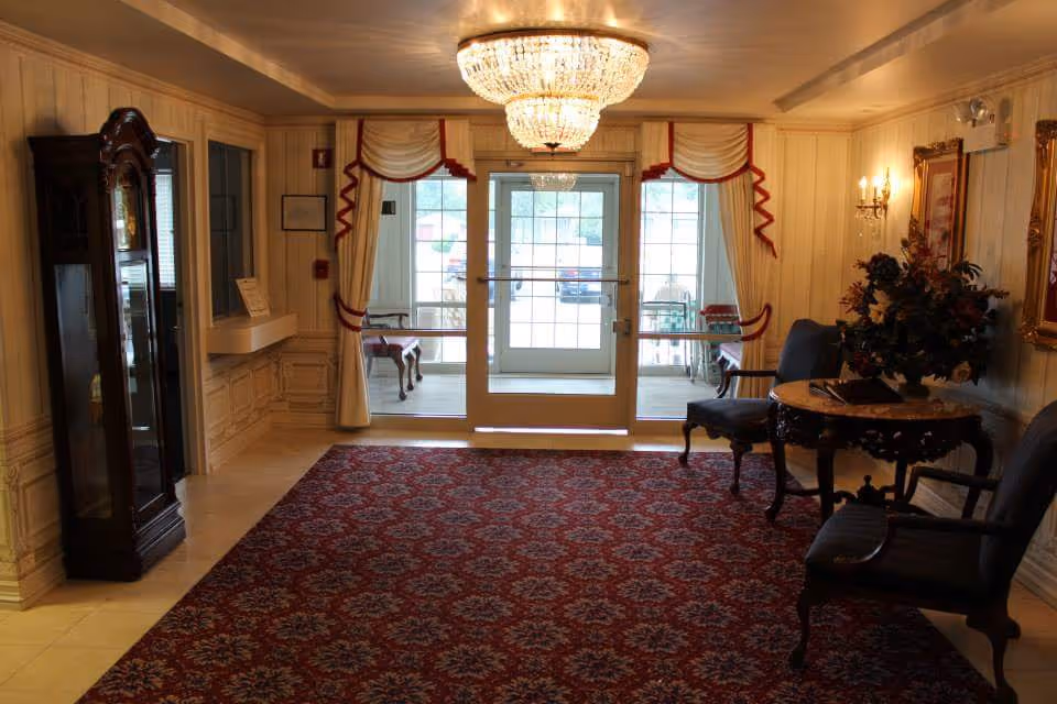 An elegant interior entryway with a large red patterned carpet, two upholstered chairs, a round wooden table with a floral arrangement, a tall grandfather clock, and a crystal chandelier hanging from the ceiling. The room has cream-colored walls with decorative molding and a glass door leading outside, flanked by windows with cream curtains trimmed in red.