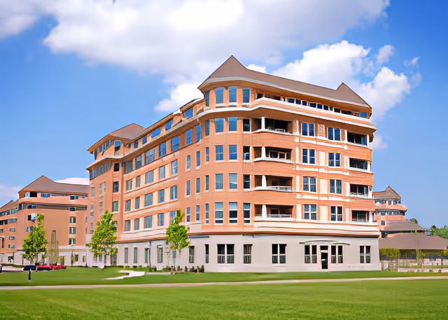 A large multi-story brick building with numerous windows and balconies, set against a blue sky with some clouds. The building is surrounded by a well-maintained green lawn and a few small trees.