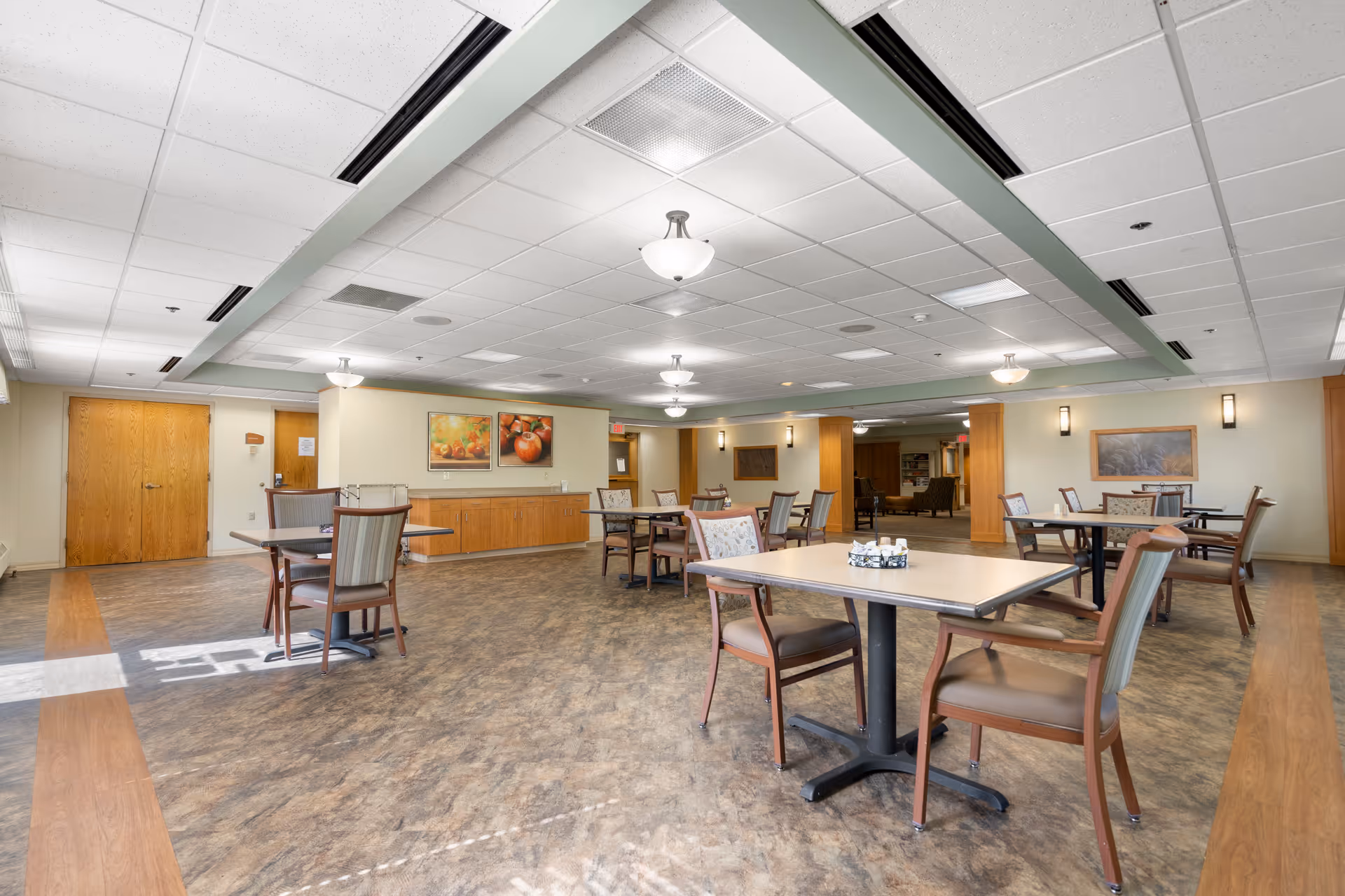 Spacious dining room with multiple tables and chairs, wood cabinetry, and framed artwork along the walls.