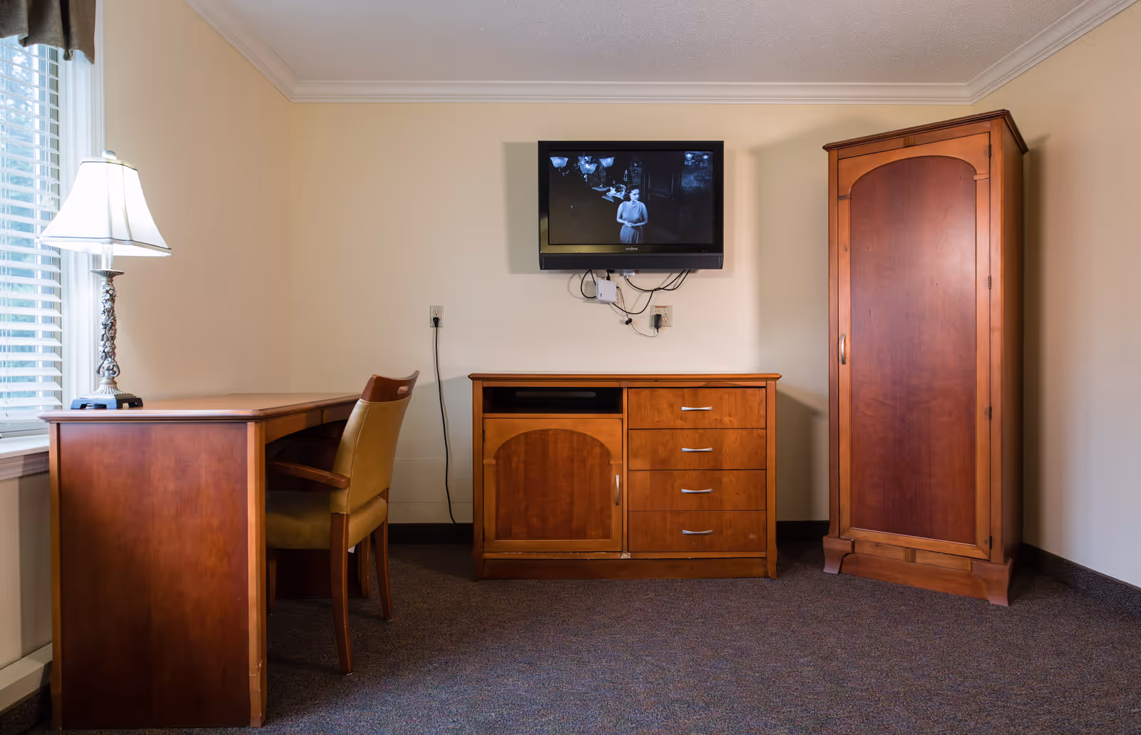 A simple furnished room with a wooden desk and chair by a window, a TV mounted above a wooden dresser, and a wooden wardrobe.