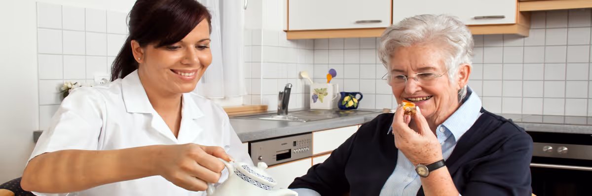 A caregiver in a white uniform pours tea from a teapot while an elderly woman with white hair and glasses smiles and eats a small pastry in a kitchen setting.