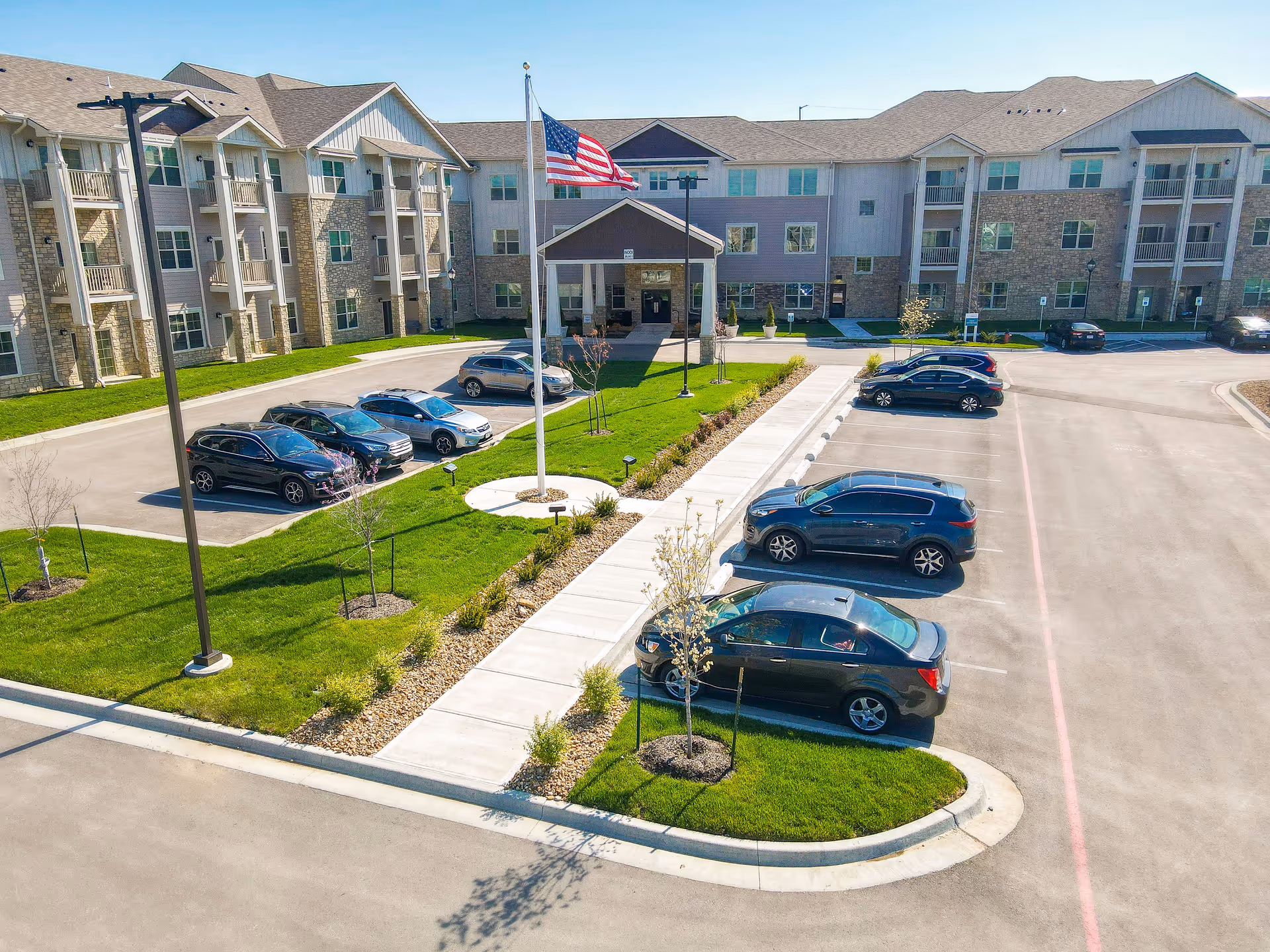 Exterior view of The Madison Senior Living facility showing a three-story building with balconies, an American flag on a flagpole, a parking lot with several cars, and landscaped green areas with small trees and shrubs.
