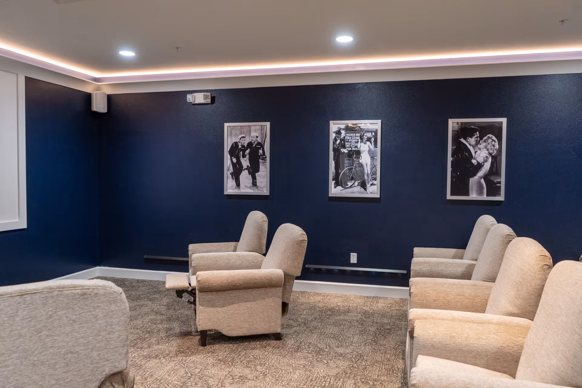 A cozy room with beige recliner chairs arranged in rows facing a dark blue wall adorned with three black and white framed photographs. The room has recessed ceiling lights and a patterned carpet floor.