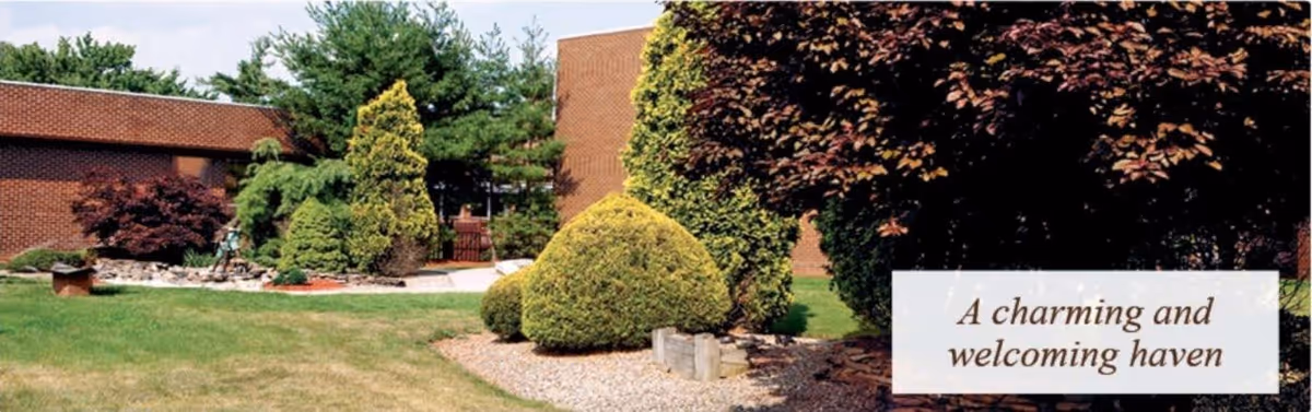 Outdoor garden area with neatly trimmed bushes, trees, and a lawn in front of a brick building. There is a small statue and a pathway visible among the greenery. A text overlay in the bottom right corner reads 'A charming and welcoming haven'.
