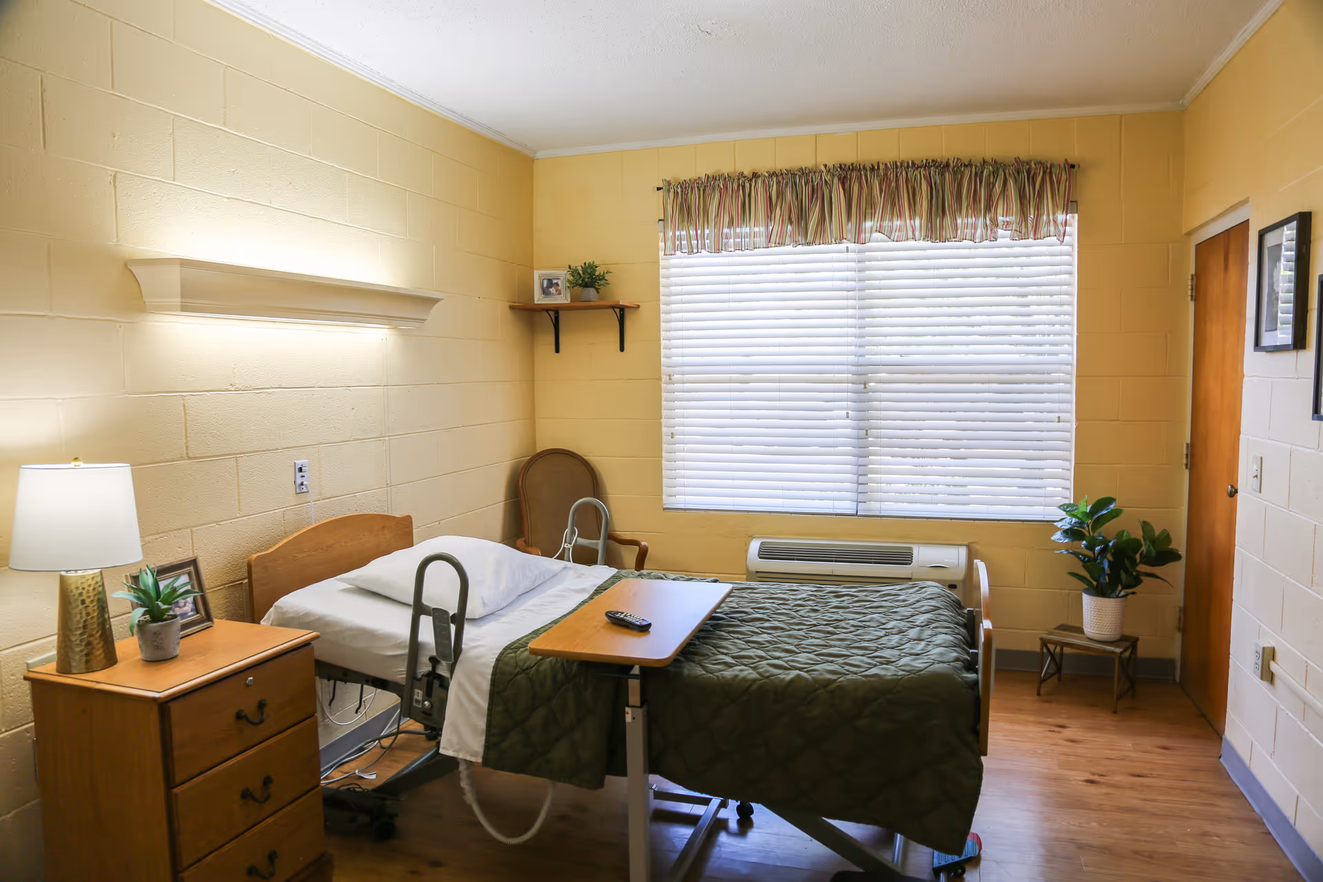 A single-occupancy senior care bedroom with a hospital bed, overbed table, nightstand with lamp, chair, and a large window with blinds.