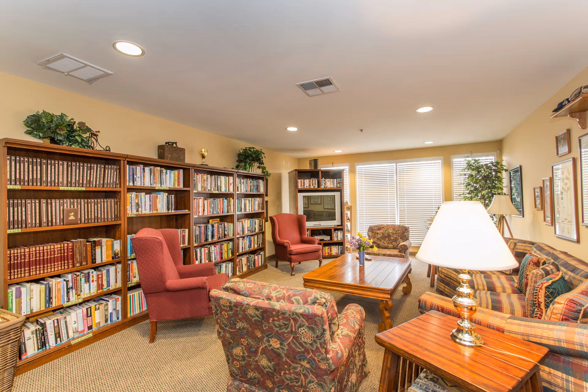 A cozy common room with bookshelves lining the wall, upholstered armchairs and sofas surrounding a wooden coffee table and a TV.