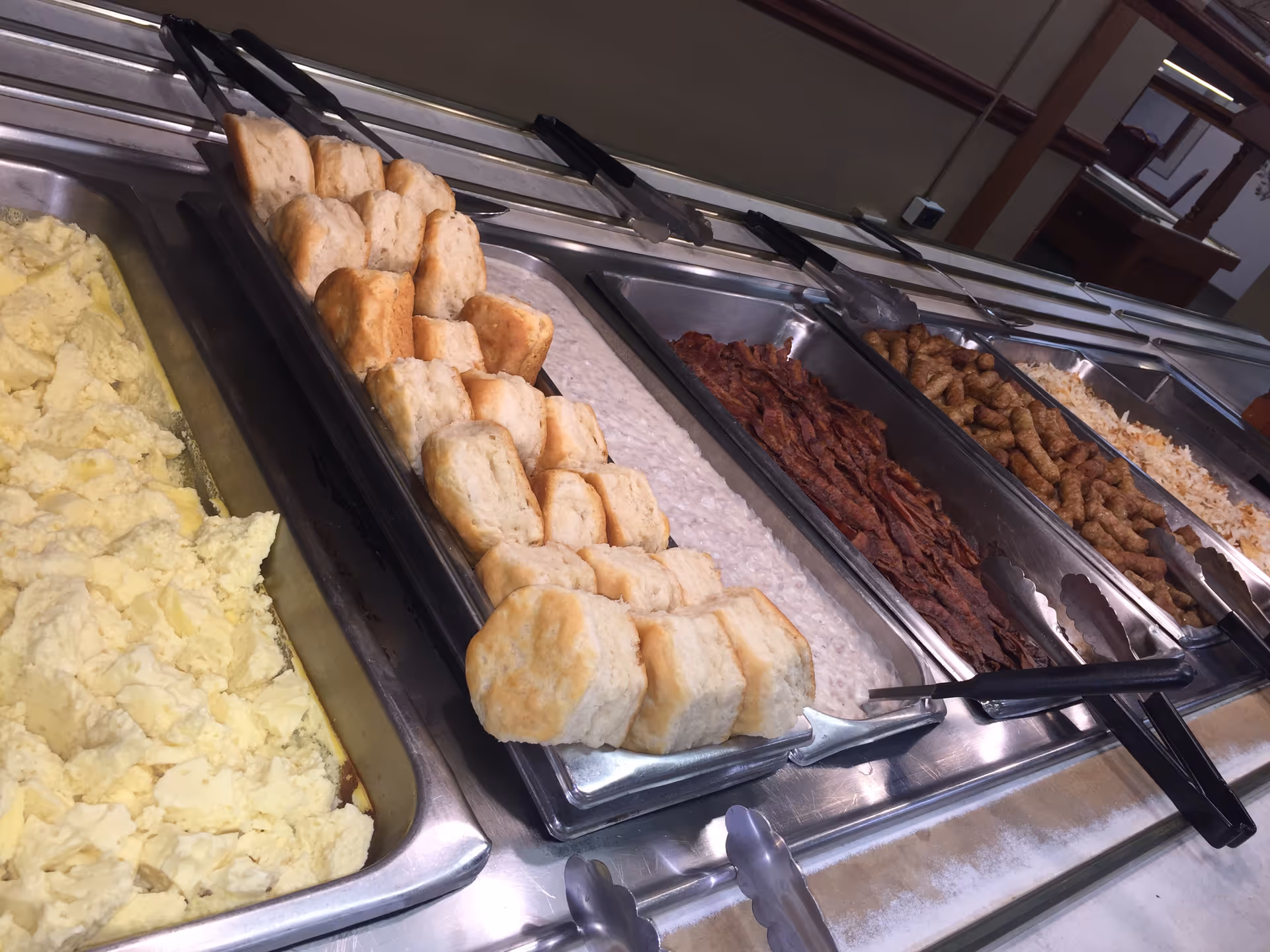A buffet-style food serving station with trays containing scrambled eggs, biscuits, gravy, bacon, sausage links, and hash browns. Serving tongs are placed on each tray.