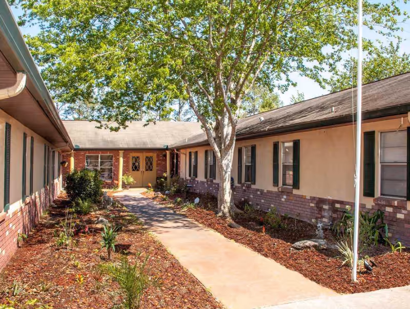 Outdoor walkway leading to the entrance of a single-story building with brick and beige walls, green window shutters, and a large tree providing shade. The walkway is bordered by landscaped garden beds with mulch and various plants.