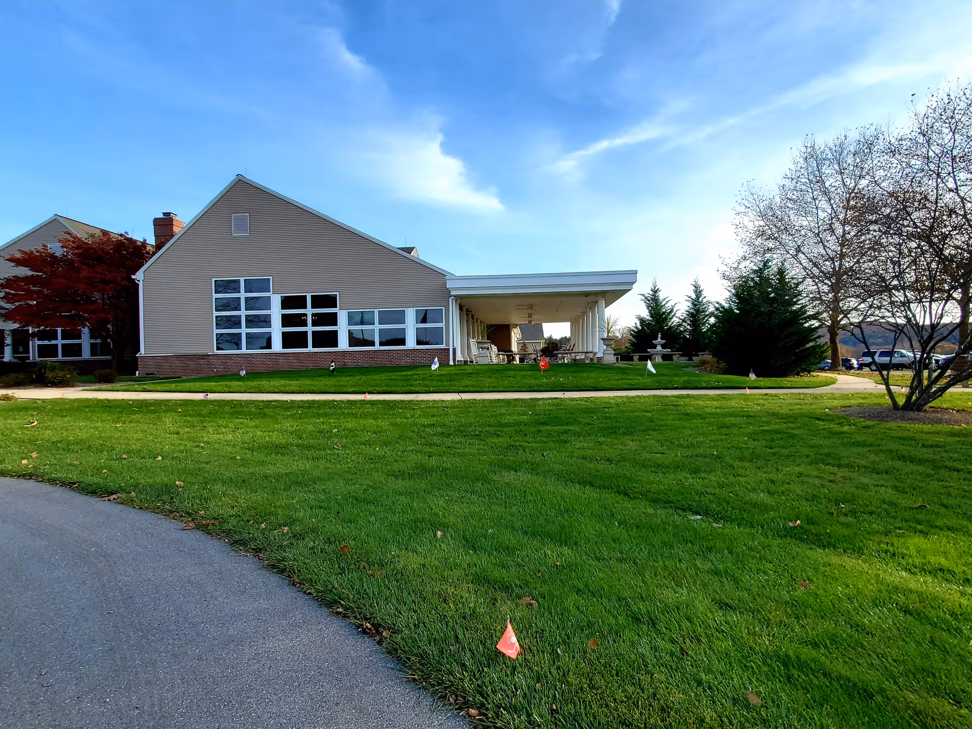 Exterior view of a single-story building with beige siding and large windows, surrounded by a well-maintained green lawn and a paved pathway. There are trees with autumn foliage and a clear blue sky with some clouds.