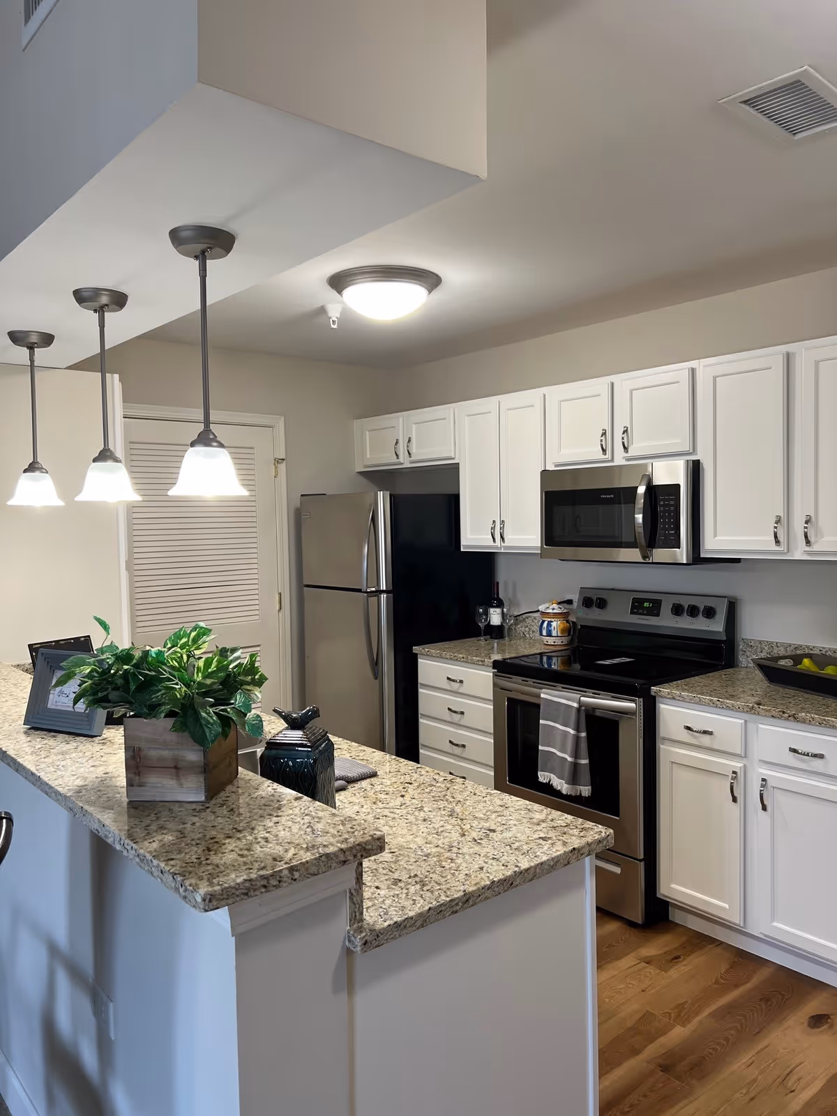 Modern kitchen with white cabinets, granite countertops, stainless steel refrigerator, stove, and microwave. Three pendant lights hang over the counter, which has a small potted plant and decorative items. The floor is wooden, and there is a towel hanging on the oven handle.