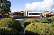 Exterior view of University Park Care Center showing a single-story building with a covered entrance and a parked shuttle bus. The foreground features neatly trimmed round green bushes and a paved driveway under a partly cloudy sky.