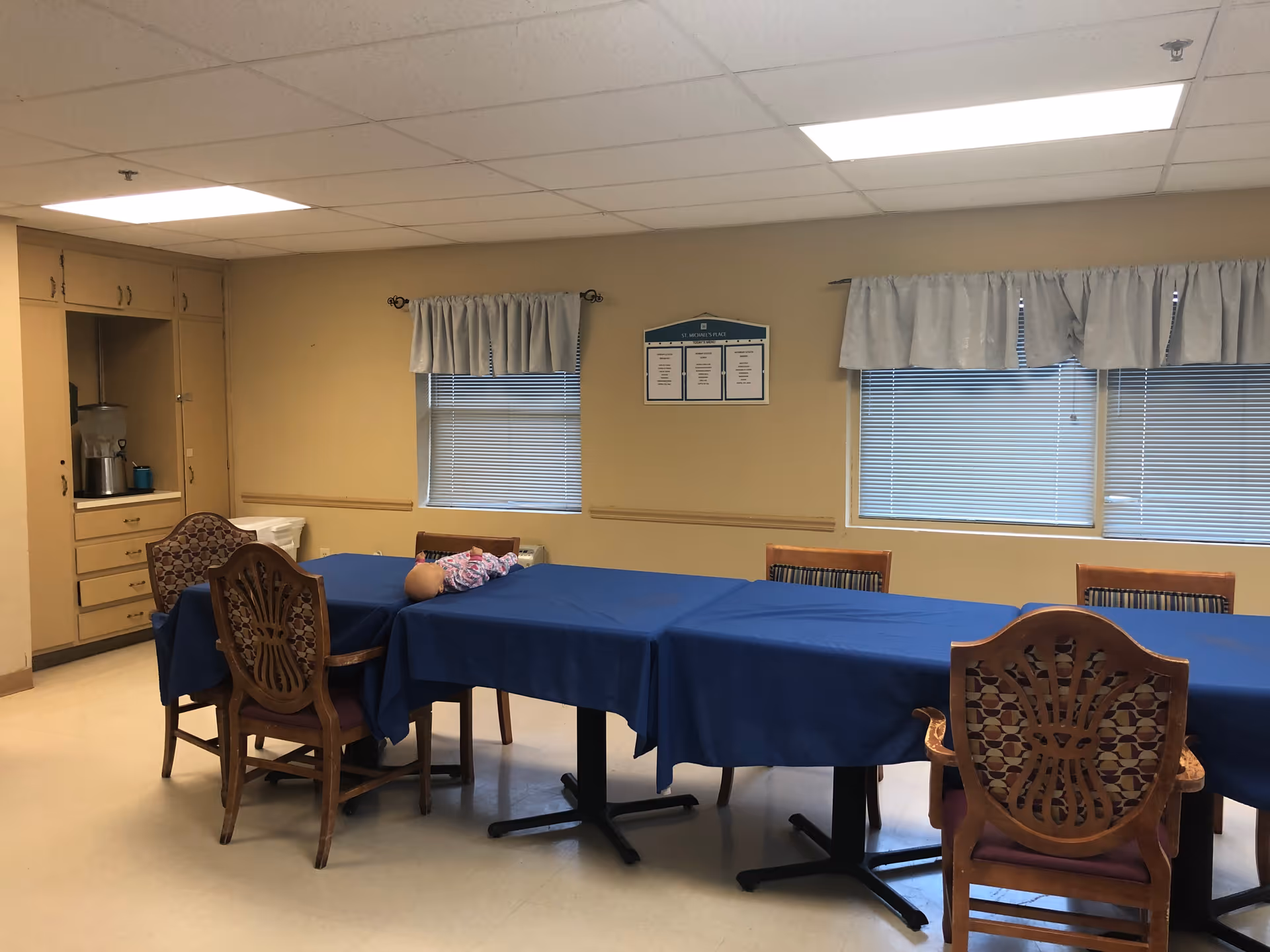 Communal dining room with blue-covered tables, wooden chairs, windows with valances, and overhead fluorescent lighting.