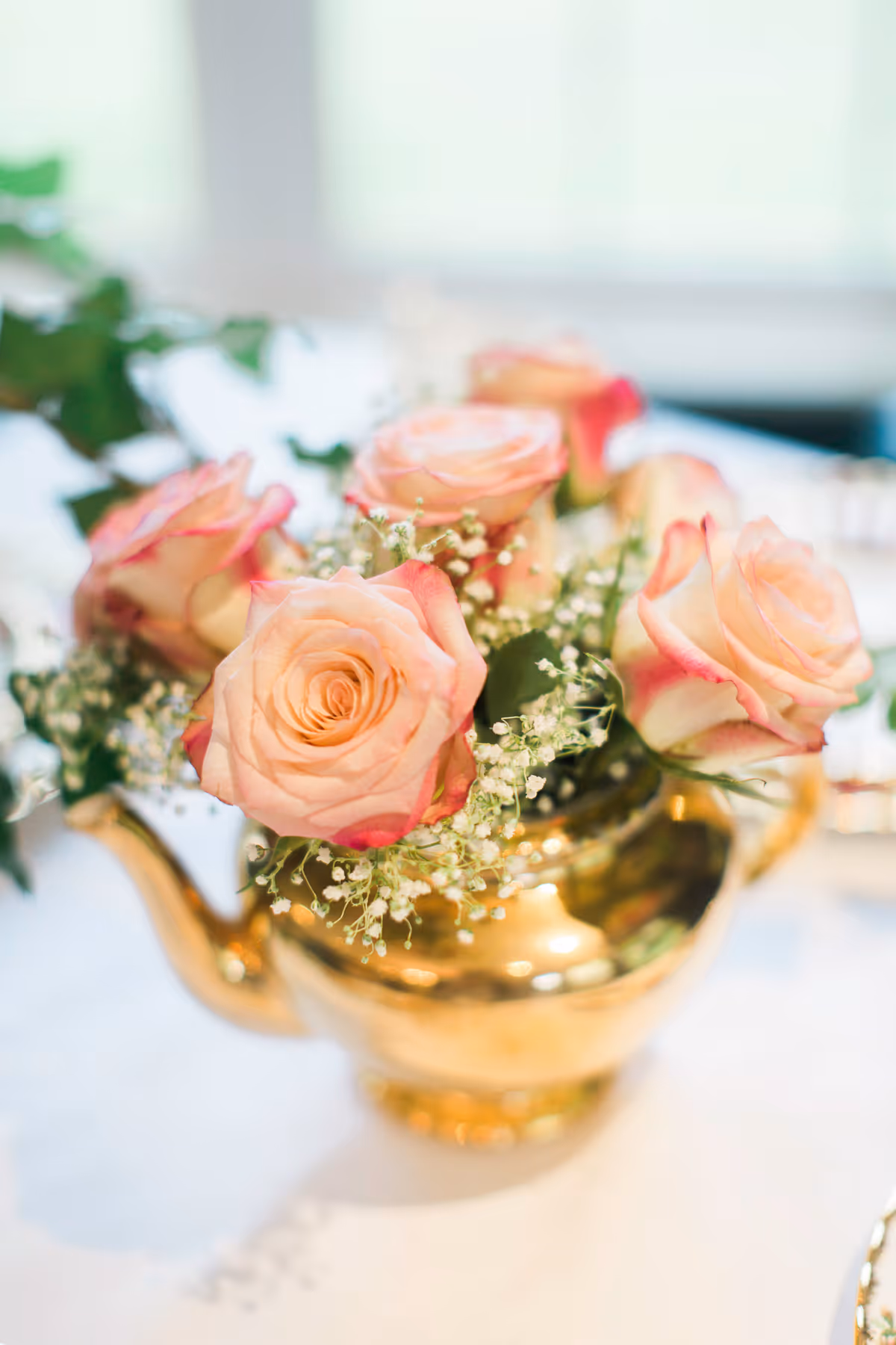 Pink roses and baby's breath arranged in a gold teapot used as a table centerpiece.