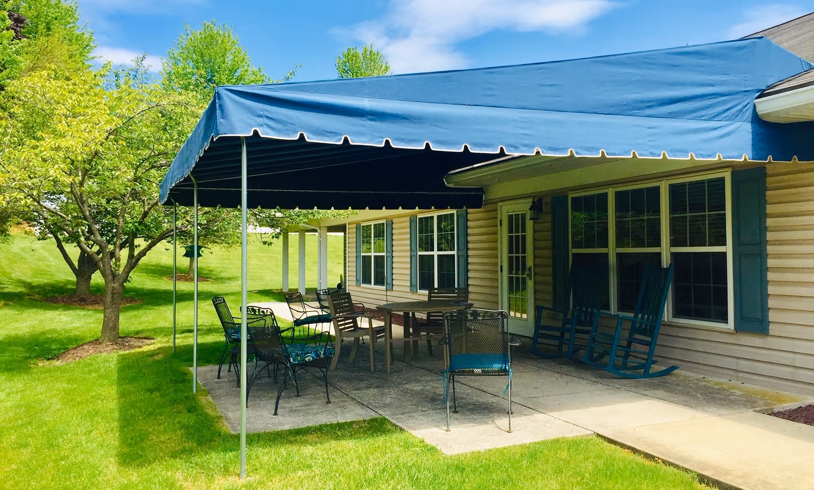 Outdoor patio area at Linden Village A ProMedica Senior Living Community with a blue canopy providing shade over several metal and wooden chairs and tables. The patio is adjacent to a beige building with blue shutters and windows, surrounded by green grass and trees under a partly cloudy sky.