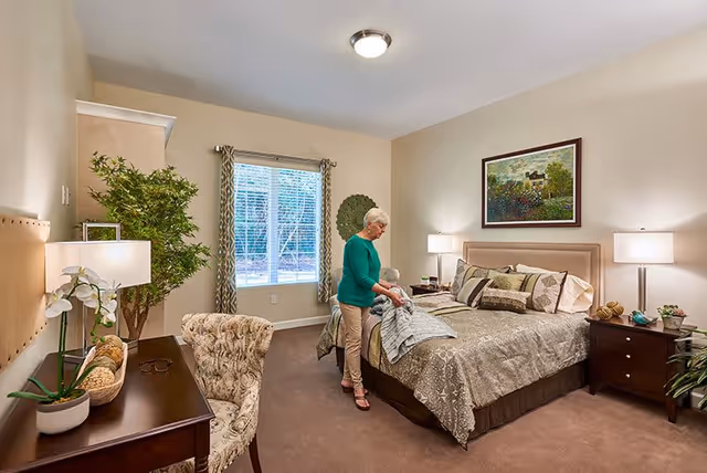 A senior woman making the bed in a well-lit bedroom with beige walls and carpet. The room features a large window with patterned curtains, a wooden desk with a chair, two bedside tables with lamps, a potted plant, and a framed painting above the bed.