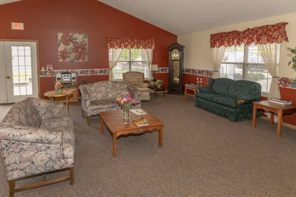 Assisted living common room with multiple upholstered sofas and armchairs arranged around a wooden coffee table, windows, and a grandfather clock.