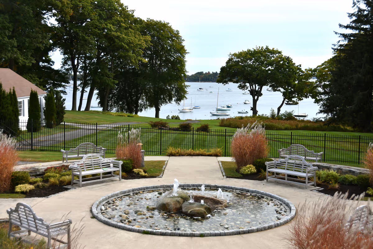 Outdoor garden area with a circular stone fountain in the center surrounded by benches and ornamental grasses. Beyond the garden is a grassy lawn with trees and a view of a body of water with several sailboats anchored.