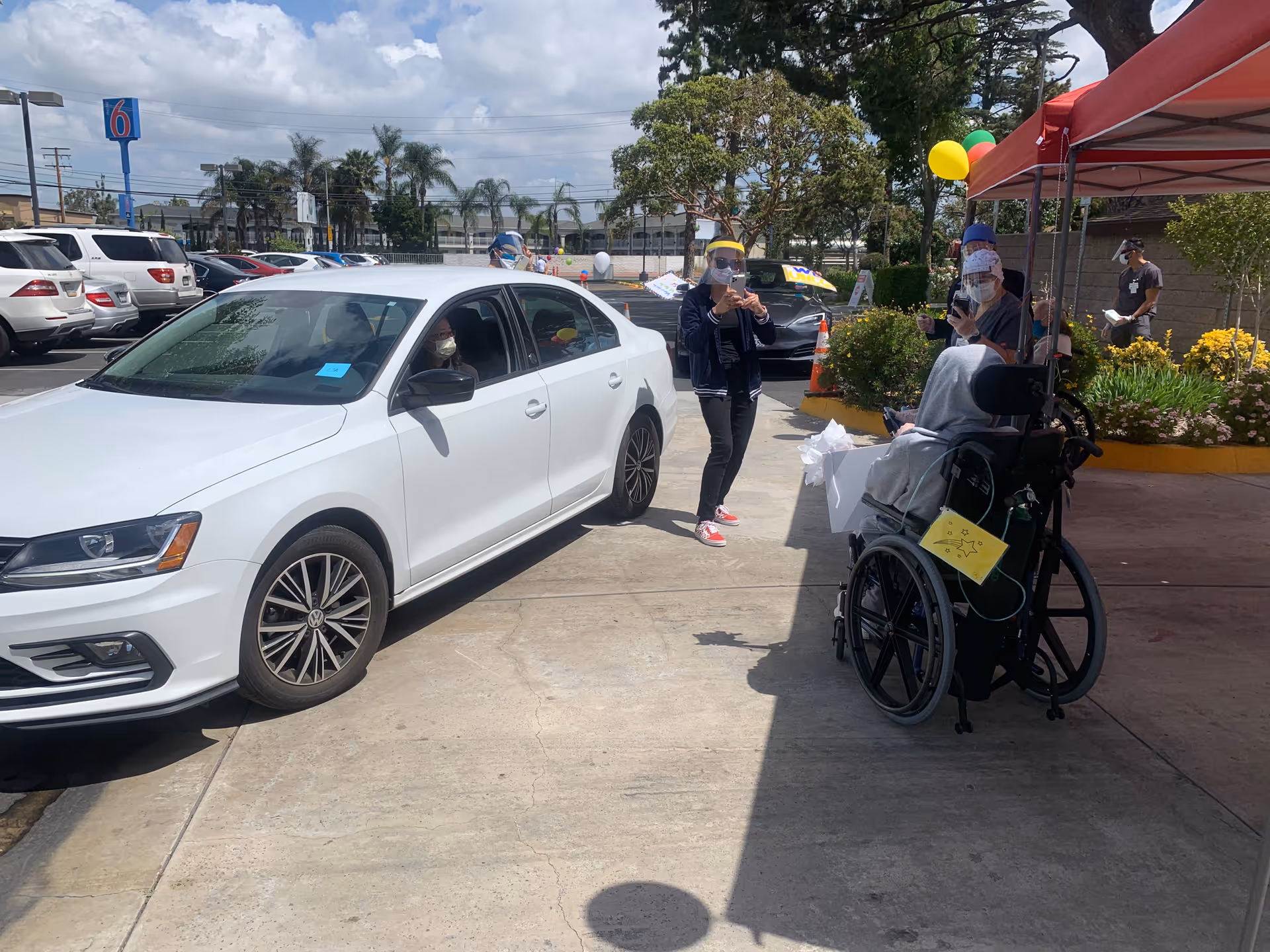 A white sedan in a facility driveway with staff wearing face shields interacting with a person in a wheelchair under a canopy.