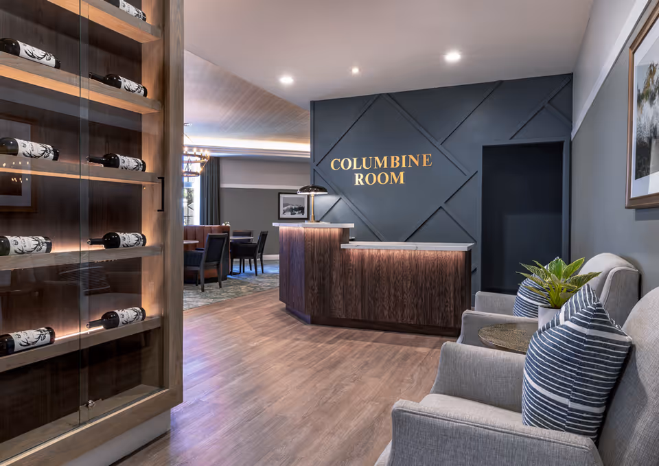 Interior view of a senior living facility's Columbine Room entrance area featuring a wooden reception desk, a dark accent wall with gold lettering, a glass cabinet displaying wine bottles, and a seating area with two gray armchairs and striped pillows next to a small table with a plant.