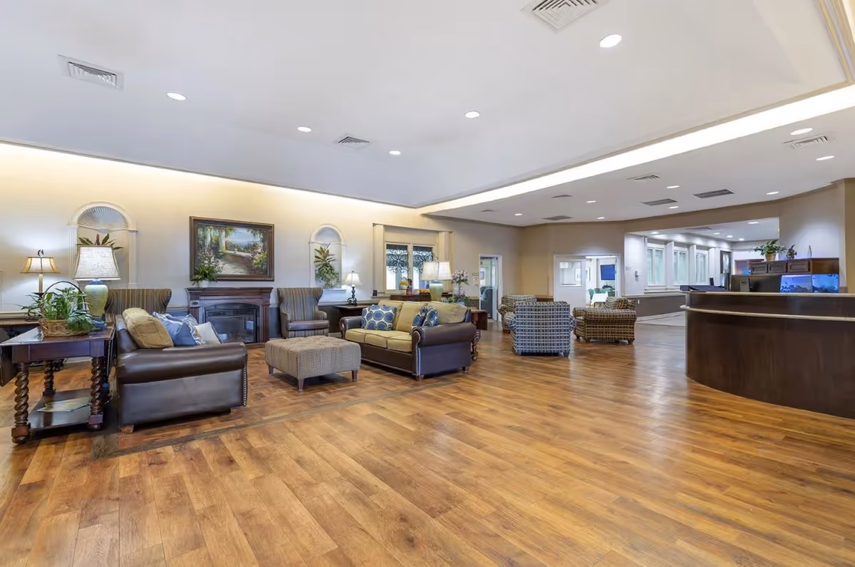 Spacious lounge area in a senior living facility with sofas, armchairs, a fireplace, side tables, and a reception desk on wood floors.