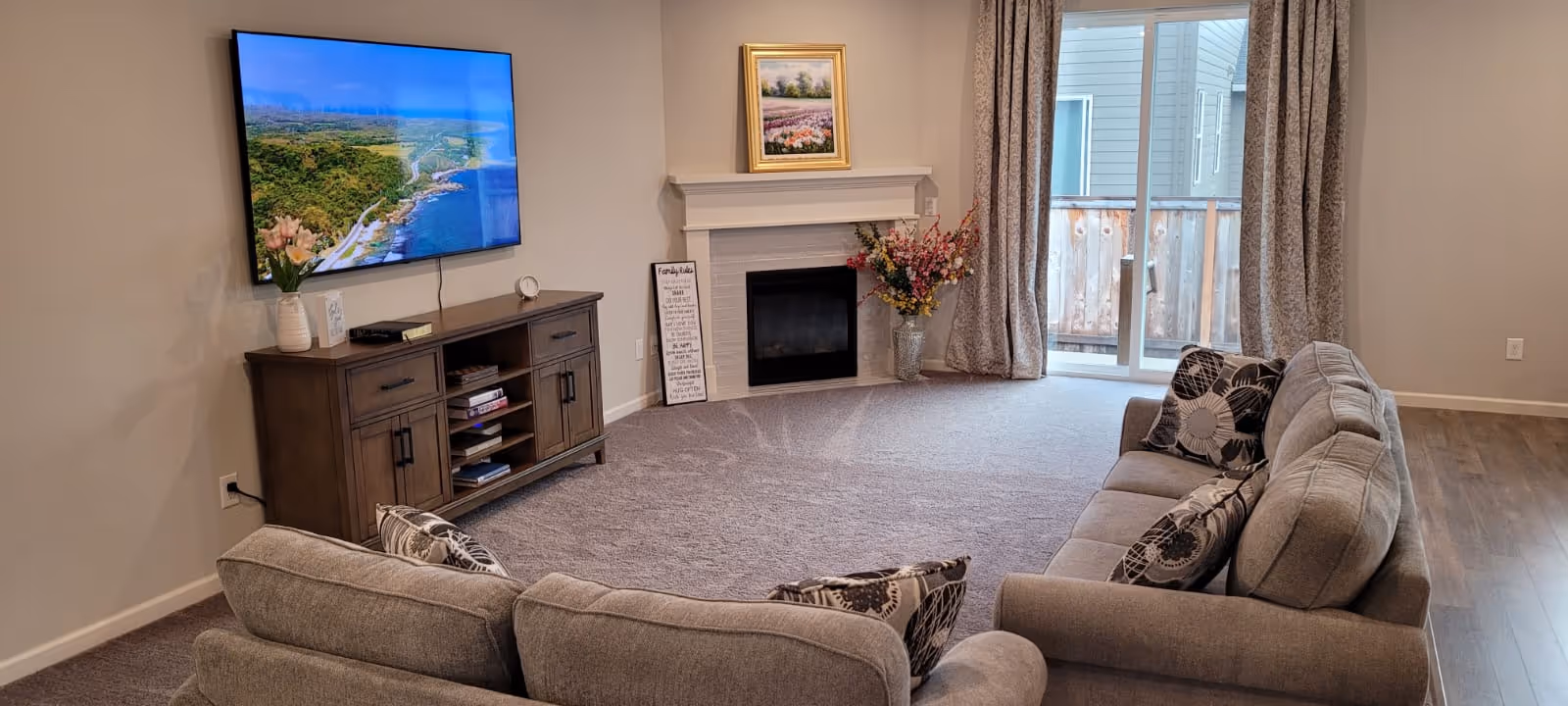 A cozy living room with a large gray sectional sofa adorned with patterned pillows. A flat-screen TV is mounted on the wall above a wooden media console. There is a white fireplace with a framed painting above it and a vase with flowers beside it. Sliding glass doors with curtains lead to an outdoor area with a wooden fence.
