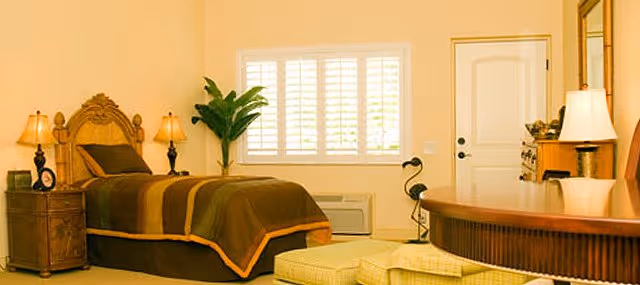 Sunlit bedroom with a single bed, nightstands and lamps, a dresser and mirror, window with shutters, and an ottoman.