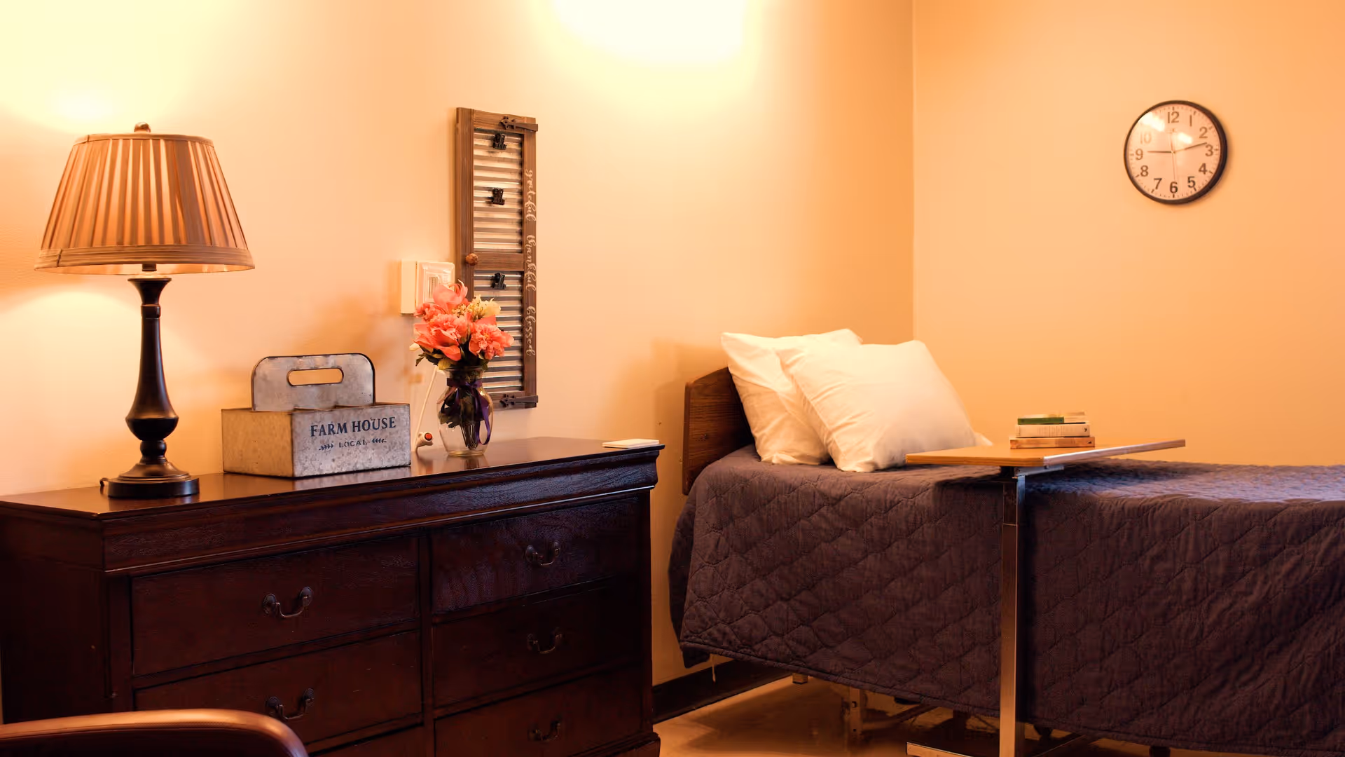 A cozy bedroom with a single bed covered in a dark quilt, two white pillows, and a small overbed table with books on it. Next to the bed is a dark wooden dresser with a lamp, a decorative box labeled 'FARM HOUSE', and a vase of pink flowers. A clock is mounted on the beige wall above the bed.