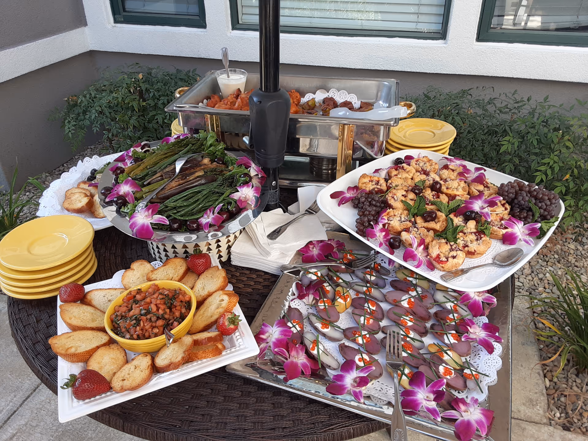 Outdoor buffet table with various appetizers including toasted bread with tomato topping, grilled vegetables, mini muffins with grapes and mint, and small hors d'oeuvres garnished with purple flowers. Yellow plates and napkins are stacked nearby, with greenery and a window in the background.