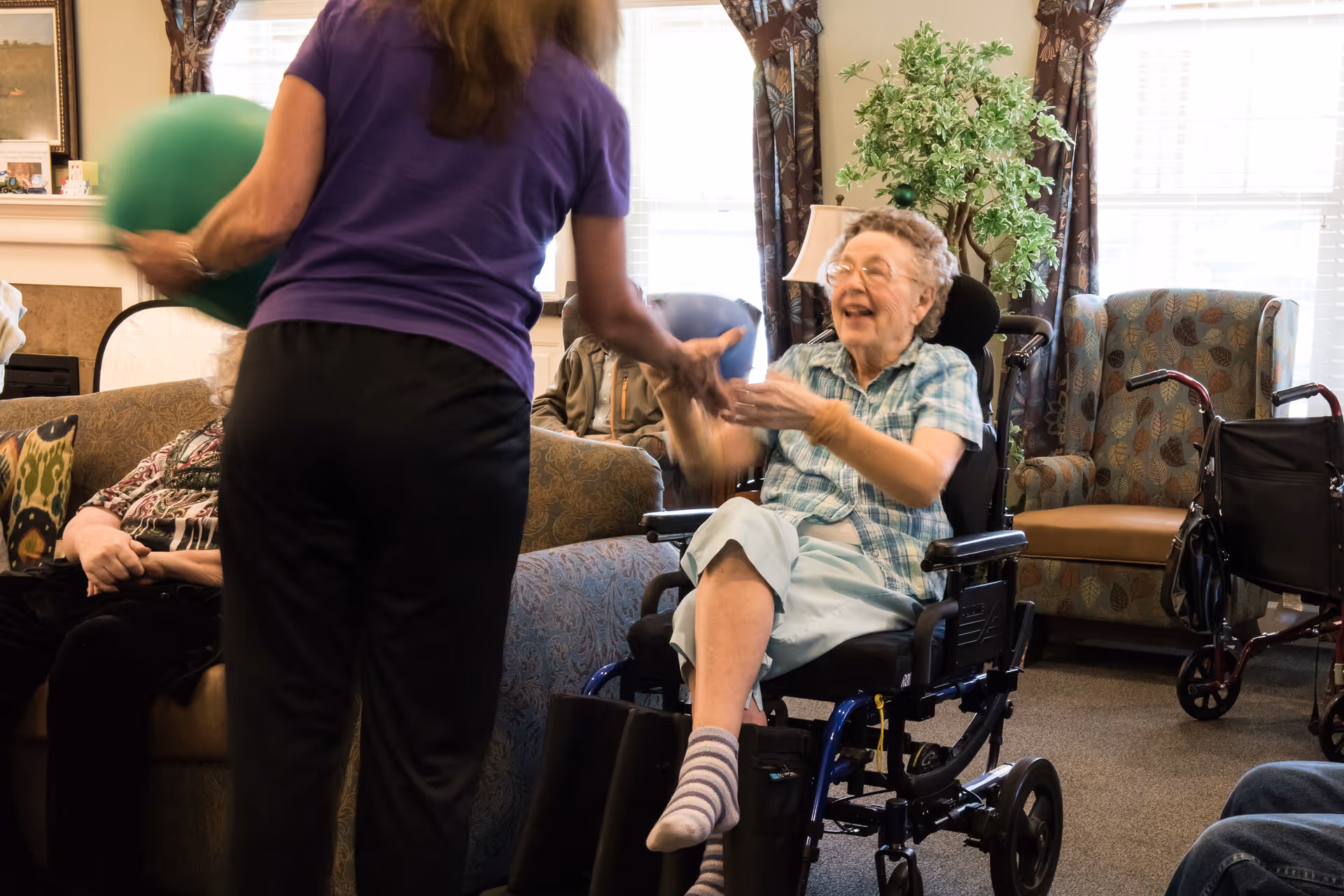 An elderly woman in a wheelchair smiling and reaching out to catch a green ball being thrown by a standing woman in a purple shirt inside a cozy living room with armchairs, a sofa, and a potted plant near the window.