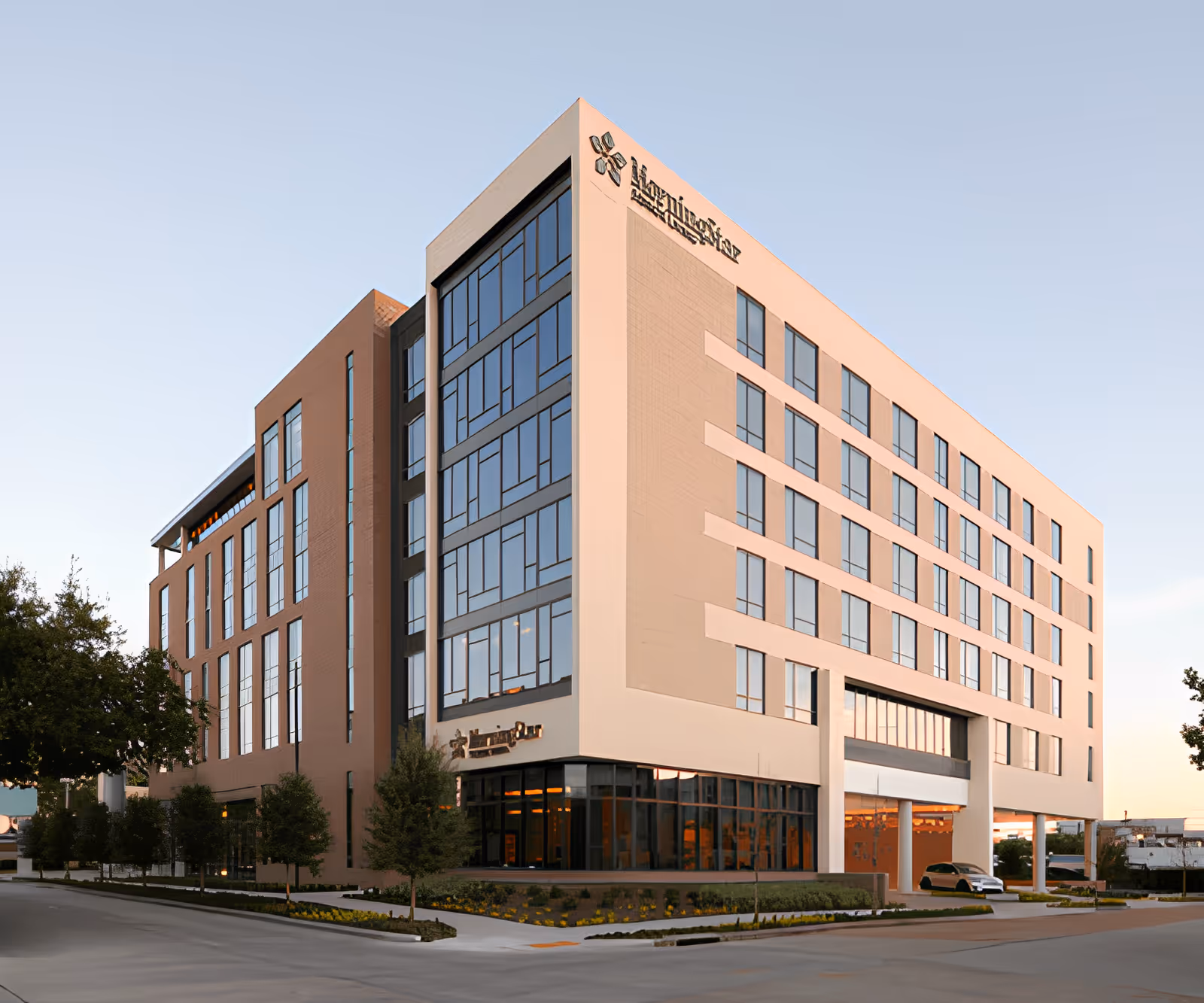 Exterior view of a modern multi-story building with large windows and a combination of light beige and brown brick facade, identified as MorningStar Senior Living at River Oaks, surrounded by landscaped greenery and a clear sky.