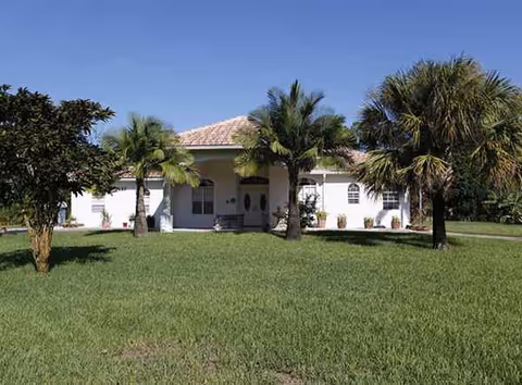Single-story white facility with tile roof, palm trees, and a grassy front lawn.