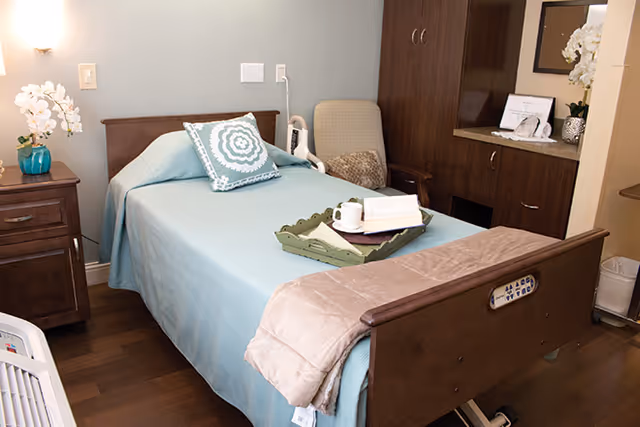 A neatly made single bed with a light blue bedspread and decorative pillow in a senior living facility room. There is a wooden nightstand with a vase of white flowers on the left, a beige armchair next to the bed, and a wooden cabinet with a countertop and decorative items in the background. A tray with a cup and an open book rests on the bed.