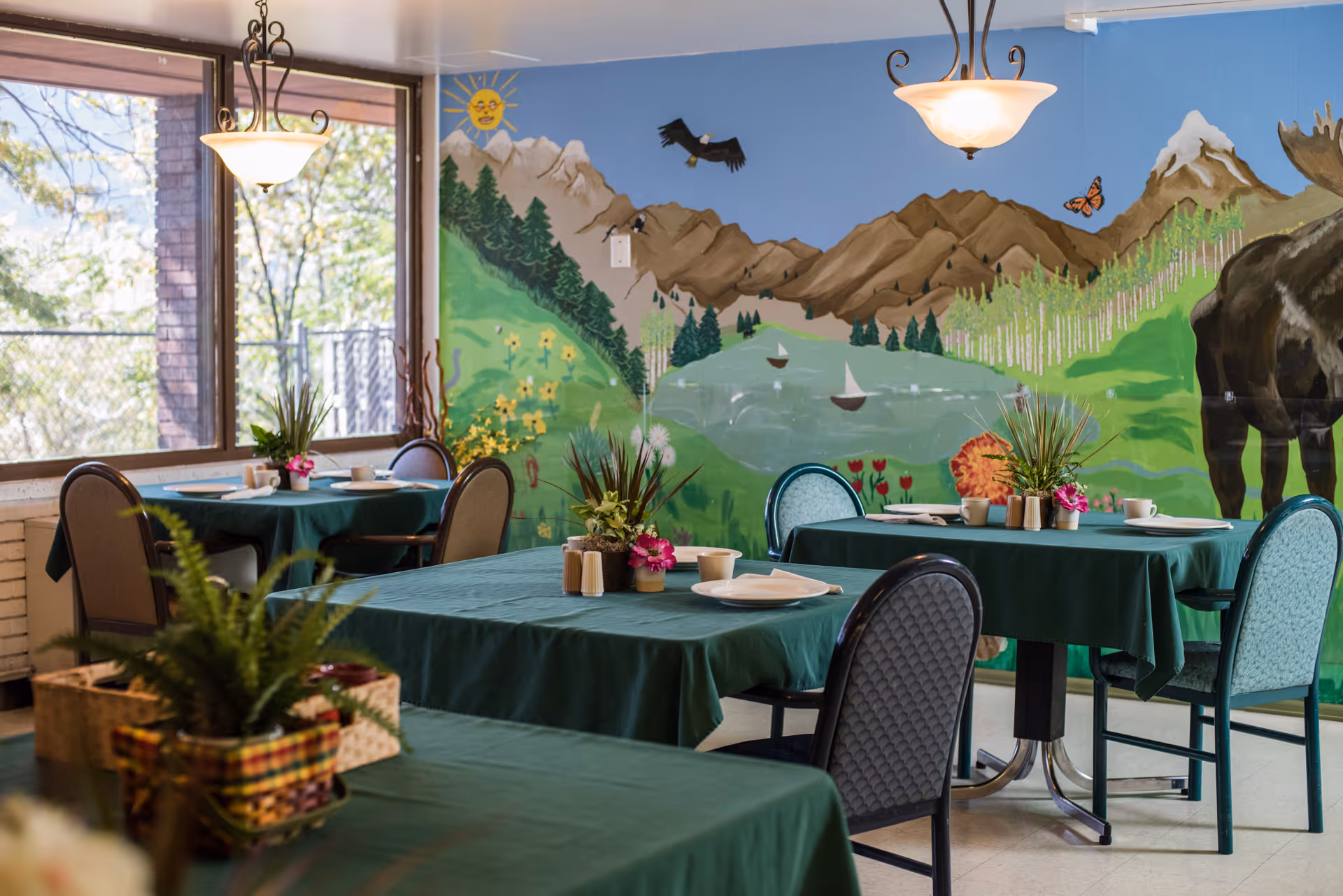 Dining room with green tablecloths, chairs, potted centerpieces, large windows, and a painted mountain mural on the wall.