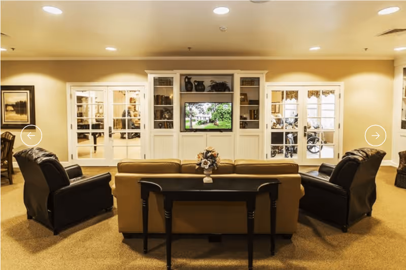 A cozy living room area in an assisted living facility featuring a beige sofa with a black console table behind it, two black leather armchairs, a built-in white entertainment center with shelves and a TV displaying a garden scene, and glass-paneled double doors on either side of the entertainment center. The room has warm lighting and beige carpeting.